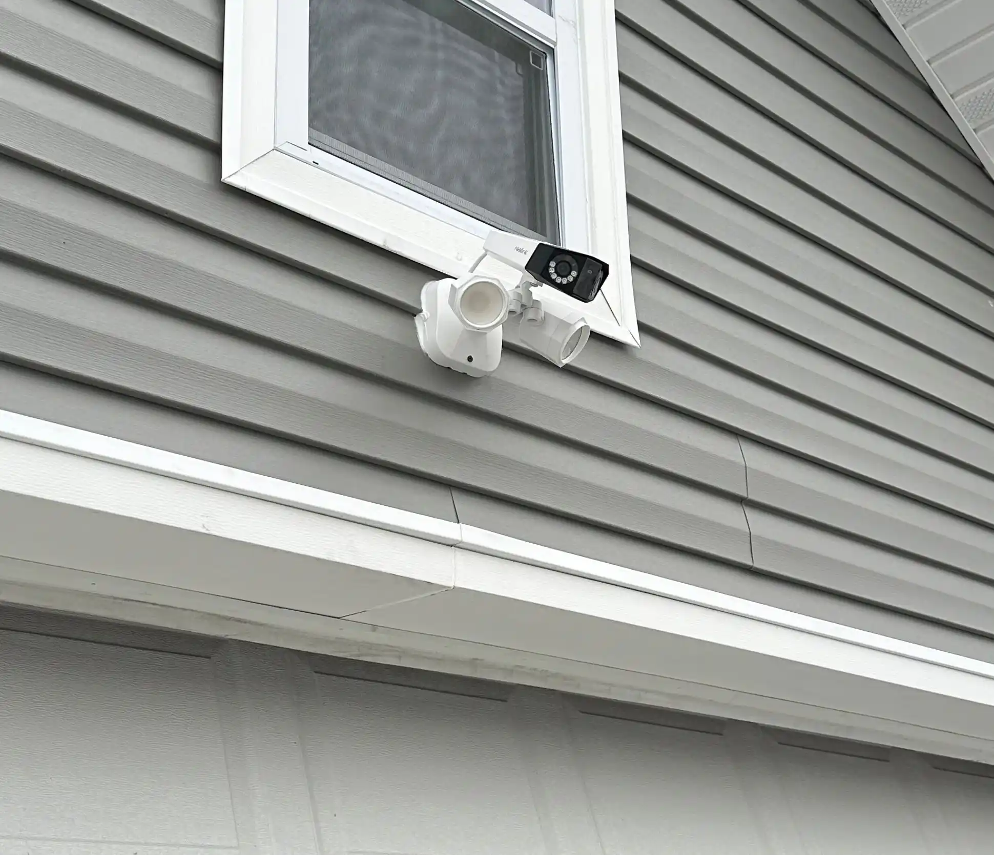 Bullet security camera and floodlight mounted on residential gray siding above garage
